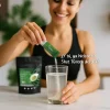 Alt Text (Imagen Principal): Mujer joven sonriente preparando su Shot T-Green de Health Addiction en un vaso de agua, representando un ritual de bienestar diario.
