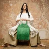 Modelo vestida en tonos neutros sostiene un bolso artesanal verde de crochet con flecos, pieza de autor de María Mulata, fotografiada frente a un muro texturizado en un entorno editorial.