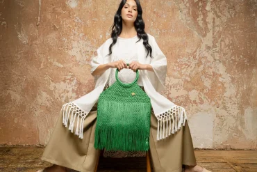 Modelo vestida en tonos neutros sostiene un bolso artesanal verde de crochet con flecos, pieza de autor de María Mulata, fotografiada frente a un muro texturizado en un entorno editorial.