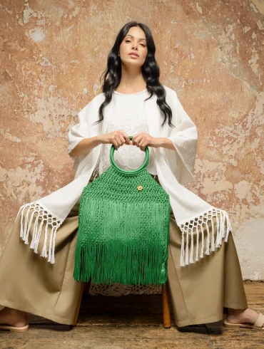 Modelo vestida en tonos neutros sostiene un bolso artesanal verde de crochet con flecos, pieza de autor de María Mulata, fotografiada frente a un muro texturizado en un entorno editorial.
