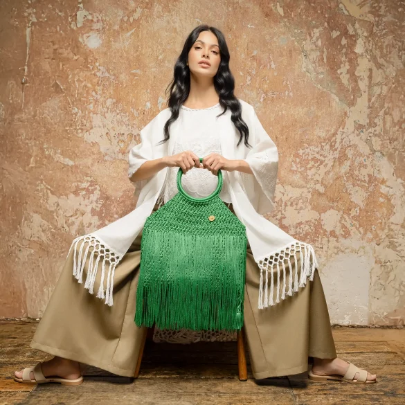 Modelo vestida en tonos neutros sostiene un bolso artesanal verde de crochet con flecos, pieza de autor de María Mulata, fotografiada frente a un muro texturizado en un entorno editorial.