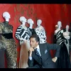 El legendario diseñador Valentino Garavani posa junto a maniquíes que lucen sus diseños de alta costura, destacando patrones de animal print y elegancia clásica frente a un icónico fondo rojo.
