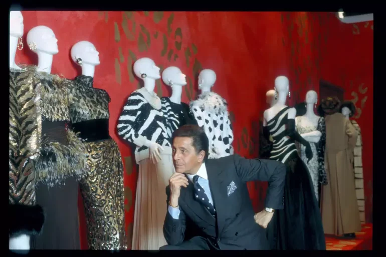 El legendario diseñador Valentino Garavani posa junto a maniquíes que lucen sus diseños de alta costura, destacando patrones de animal print y elegancia clásica frente a un icónico fondo rojo.