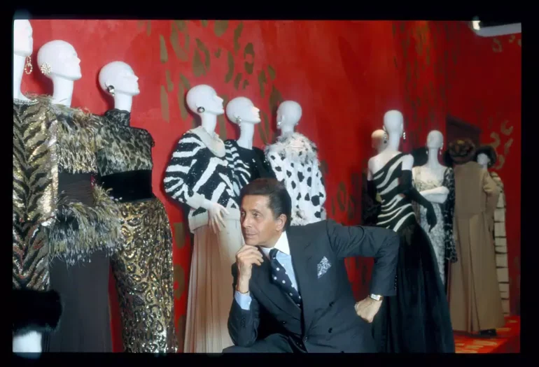 El legendario diseñador Valentino Garavani posa junto a maniquíes que lucen sus diseños de alta costura, destacando patrones de animal print y elegancia clásica frente a un icónico fondo rojo.