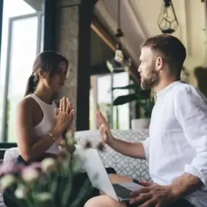 Hombre y mujer conversan frente a frente en un sofá, él con una laptop sobre las piernas y ella con las manos juntas en gesto de diálogo, en un ambiente interior con luz natural.