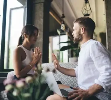 Hombre y mujer conversan frente a frente en un sofá, él con una laptop sobre las piernas y ella con las manos juntas en gesto de diálogo, en un ambiente interior con luz natural.